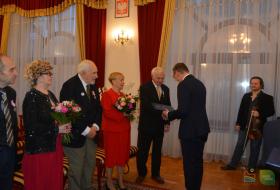 Prezydent Olsztyna wręcza jubilatowi list gratulacyjny. Po lewej stronie stoją dwie pary jubilatów, panie trzymają bukiety kwiatów, po prawej widoczny skrzypaek trzymający skrzypce. 