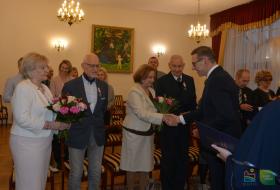 Prezydent Olsztyna wręcza jubilatce list gratulacyjny, po lewej i prawej stronie zdjęcia stoją pozostali jubilaci, w tle uczestnicy uroczystości.