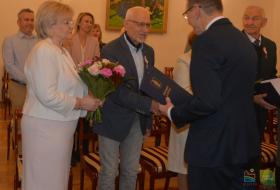 Na pierwszym planie prezydent Olsztyna wręcza list gratulacyjny jubilatowi, obok stoi jubilatka z bukietem kwiatów, w tle uczestnicy uroczystości. 