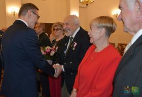 Prezydent Olsztyna gratuluje jubilatowi odznaczenia medalem za długoletnie pożycie małżeńskie. Obok stoją pozostali jubilaci.