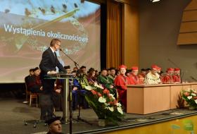 Zdjęcie przedstawia prezydenta Olsztyna stojącego przy mównicy, który zwraca się do siedzących: rektora i senatu UWM