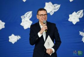 Piotr Grzymowicz, prezydent Olsztyna stoi na tle okolicznościowej dekoracji i mówi do mikrofonu