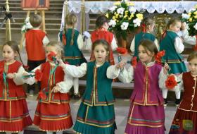 Na pierwszym planie dziewczynki w  ludowych strojach warmińskich, z wzniesionymi ramionami, tańczą przed ołtarzem, podając sobie róże