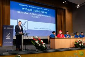 Piotr Grzymowicz, prezydent Olsztyna stoi przy mównicy podczas wystąpienia na inauguracji roku akademickiego 2022/2023 na wydziale geoinżynierii UWM. W tle ekran oraz siedzący przedstawiceiel rady wydziału.