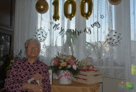 Zdjęcie przdstawia uśmiechniętą kobietę, która pozuje do zdjęcia, w tle wiszą złote balony przedstawiające liczbę sto symbolizująca 100-ne urodziny jubilatki, obok stoi wazon z bukietem kolorowych kwiatów.