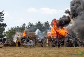 Zdjęcie przedstawia inscenizacje Bitwy pod Grunwaldem, na pierwszym planie widać rekonstruktorów, na drugim płonące chaty, w tle drzewa i niebo.