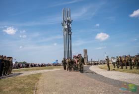 Zdjęcie przedstawia przed pomnikiem stojących żołnierzy, w tle widać dużą grupę ludzi i niebo.