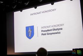 Zdjęcie przedstawia obraz wyświetlany projektorem. Na obrazie widnieje napis "Patronat Honorowy, Prezydent Olsztyna Piotr Grzymowicz" i sygnet Olsztyna.