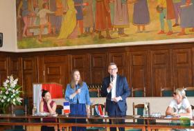 Zdjęcie przedstawia prezydenta Piotra Grzymowicza przemawiającego przez mikrofon. Razem z nim są trzy kobiety, jedna stoi, dwie siedzą przy stole.